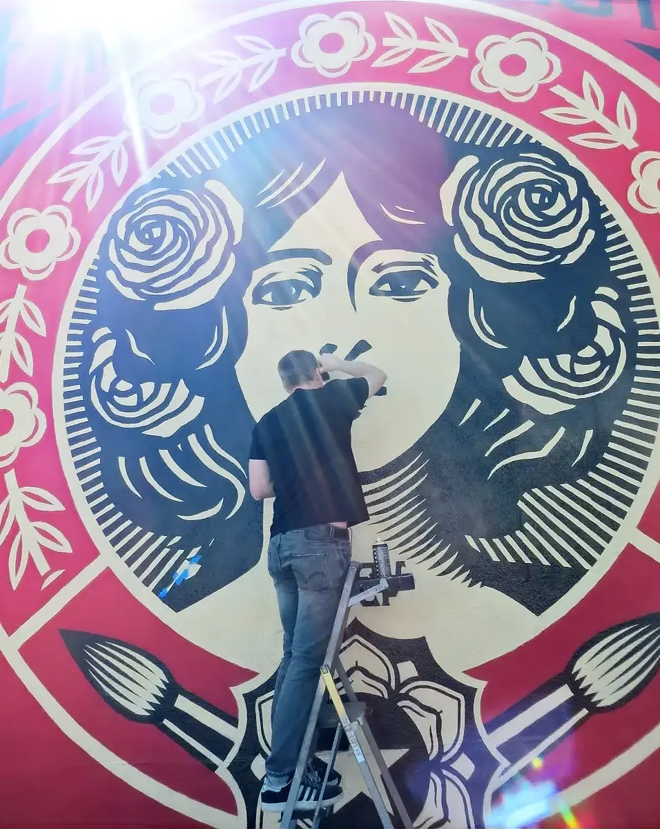 An artist on a ladder spray-painting a large red-and-cream portrait mural.